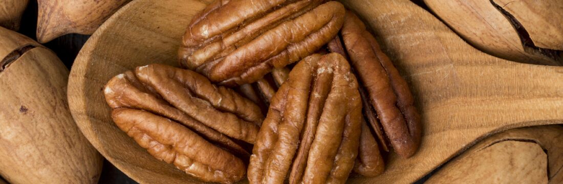 Imagine de aproape cu o lingură de lemn cu miezul unor nuci pecan, pe un fundal de nuci pecan intregi
