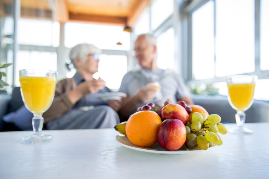In fundal cuplu in varsta care ia un mic dejun sanatos. Focus pe o farfurie cu fructe in fata lor si 2 pahare cu suc de fructe, alimente bogate in antioxidanti care sustin longevitatea